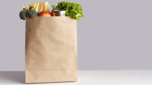 Various grocery items, including fruits and vegetables, in a paper bag on a white table opposite gray wall.